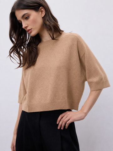 A woman wearing a cropped, short-sleeve knit sweater in a warm camel beige color, paired with black trousers, against a plain light gray studio background.