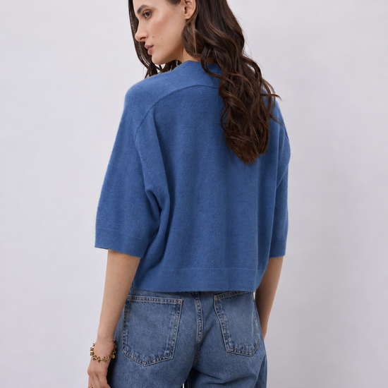 A woman shown from the back wearing a cropped, short-sleeve blue knit top and wide-leg blue jeans against a plain light gray studio background.