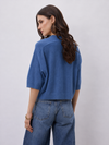 A woman shown from the back wearing a cropped, short-sleeve blue knit top and wide-leg blue jeans against a plain light gray studio background.