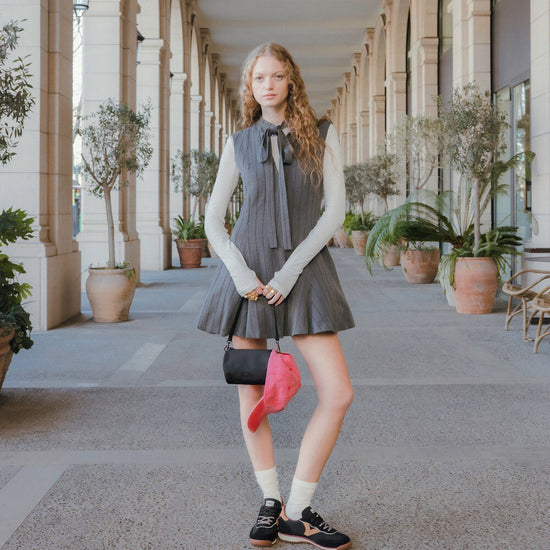 A young woman stands in a covered outdoor walkway with columns and potted plants, wearing a sleeveless pleated gray dress over a cream long-sleeve top, holding a black and pink bag, with white socks and black sneakers.