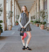 A young woman stands in a covered outdoor walkway with columns and potted plants, wearing a sleeveless pleated gray dress over a cream long-sleeve top, holding a black and pink bag, with white socks and black sneakers.