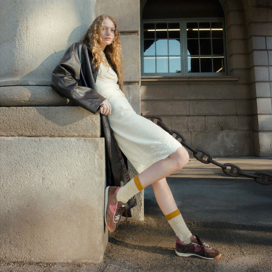 A woman leans against a stone building corner wearing a cream midi dress, a dark leather jacket, yellow socks, and brown shoes, with a large chain visible in the background.
