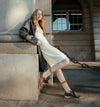 A woman leans against a stone building corner wearing a cream midi dress, a dark leather jacket, yellow socks, and brown shoes, with a large chain visible in the background.