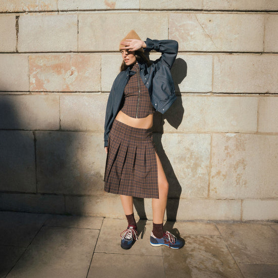 A model stands against a stone wall wearing a brown plaid crop top and matching pleated skirt set, a dark blue jacket draped off the shoulders, a tan hat, sunglasses, and blue sneakers.