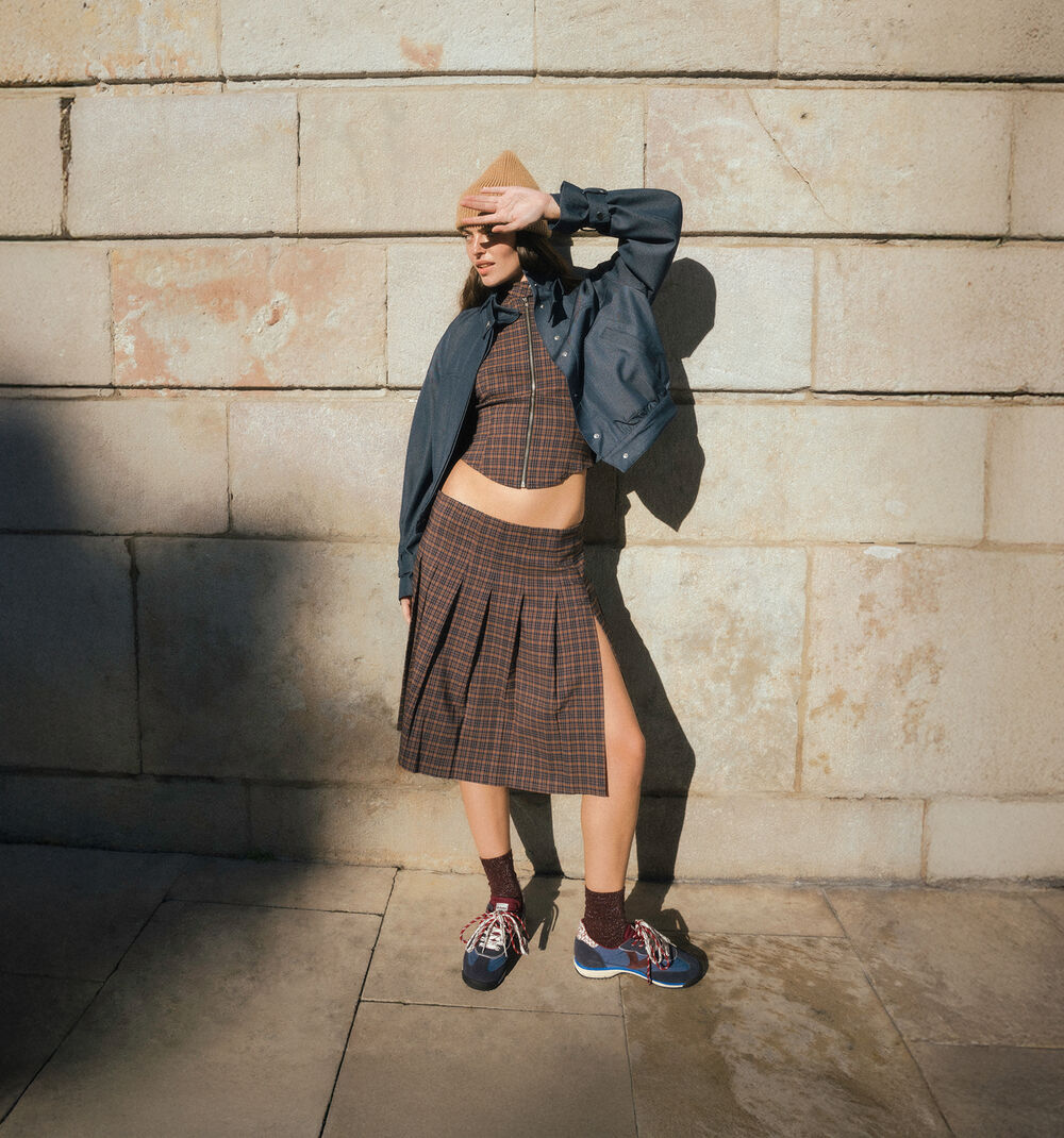 A model stands against a stone wall wearing a brown plaid crop top and matching pleated skirt set, a dark blue jacket draped off the shoulders, a tan hat, sunglasses, and blue sneakers.