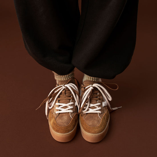 A close-up of a person's feet wearing brown suede sneakers with white laces, paired with black pants and beige socks, set against a solid dark brown background.