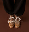 A close-up of a person's feet wearing brown suede sneakers with white laces, paired with black pants and beige socks, set against a solid dark brown background.