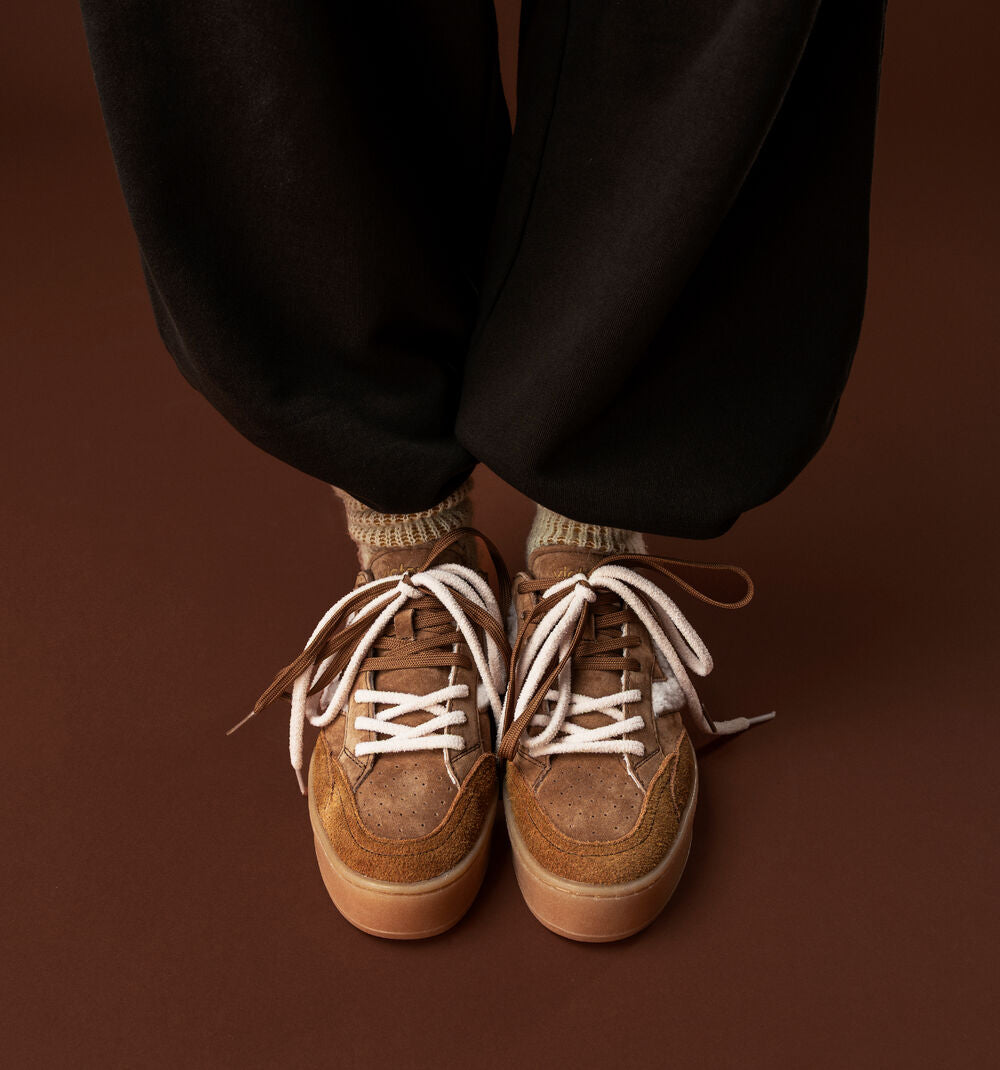 A close-up of a person's feet wearing brown suede sneakers with white laces, paired with black pants and beige socks, set against a solid dark brown background.