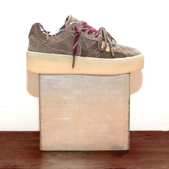 A low-top platform sneaker in taupe suede with burgundy laces, resting on a square metallic block over a wooden surface against a white background.