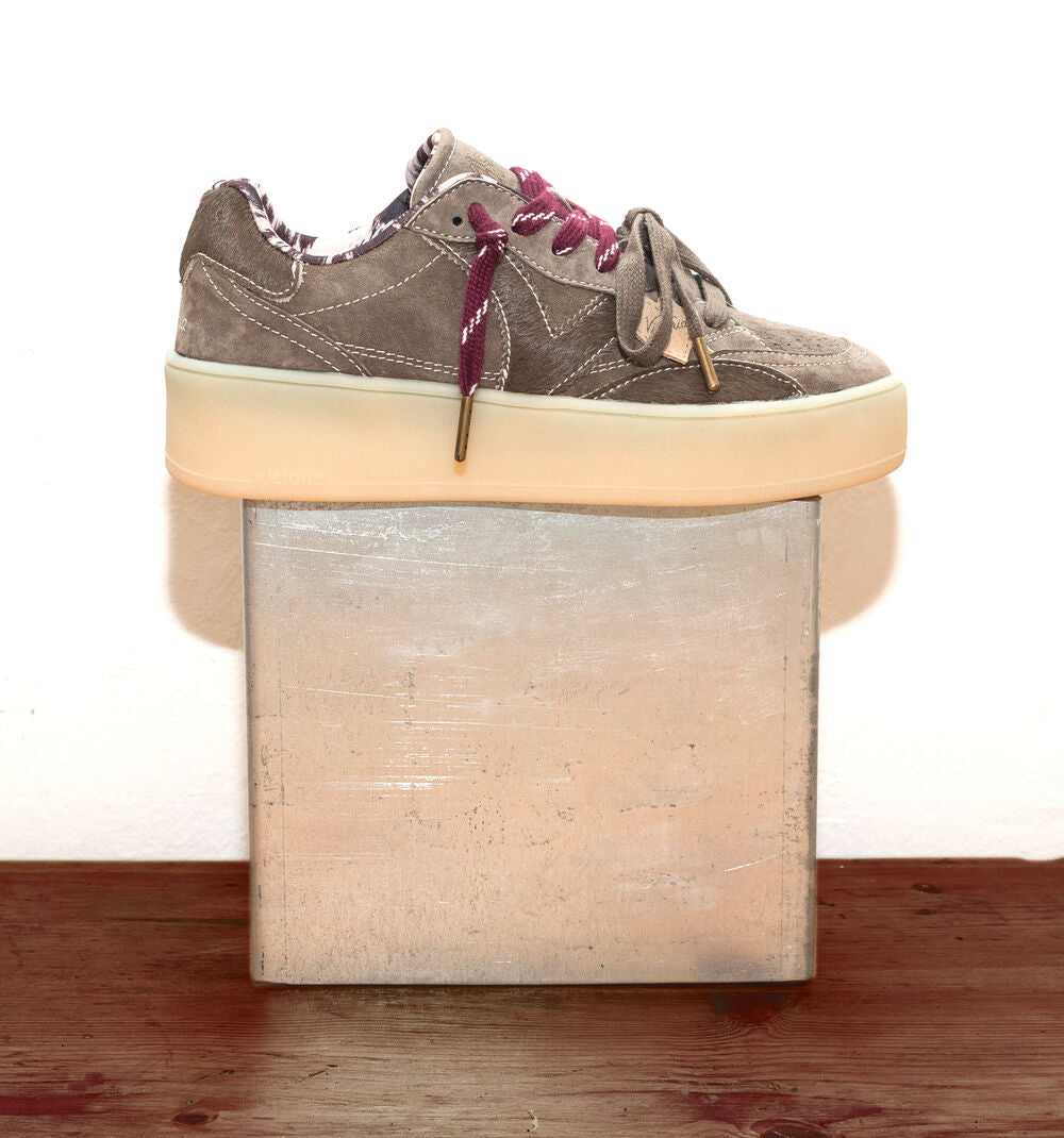 A low-top platform sneaker in taupe suede with burgundy laces, resting on a square metallic block over a wooden surface against a white background.