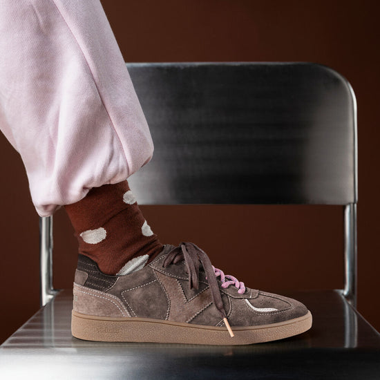 A person wearing a brown suede and canvas sneaker with pink laces, paired with a brown sock featuring white cloud-like patterns and light pink pants, sitting on a black chair against a dark brown background.