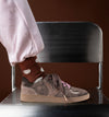 A person wearing a brown suede and canvas sneaker with pink laces, paired with a brown sock featuring white cloud-like patterns and light pink pants, sitting on a black chair against a dark brown background.