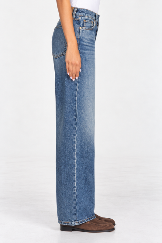 A side profile view of a person wearing wide-leg blue denim jeans and brown suede mules against a plain white studio background.