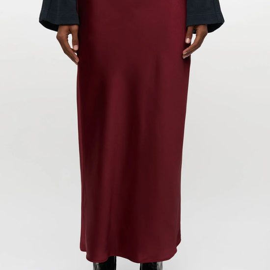 A model is shown from the waist down wearing a long, dark red satin midi skirt, a dark navy long-sleeve top, and black boots against a plain light gray background.