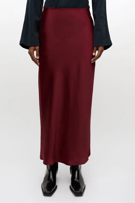 A model is shown from the waist down wearing a long, dark red satin midi skirt, a dark navy long-sleeve top, and black boots against a plain light gray background.