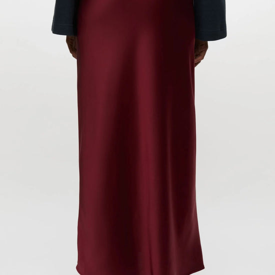A person shown from the back wearing a long satin-like burgundy skirt with a dark navy top and black shoes, photographed against a plain light gray studio background.