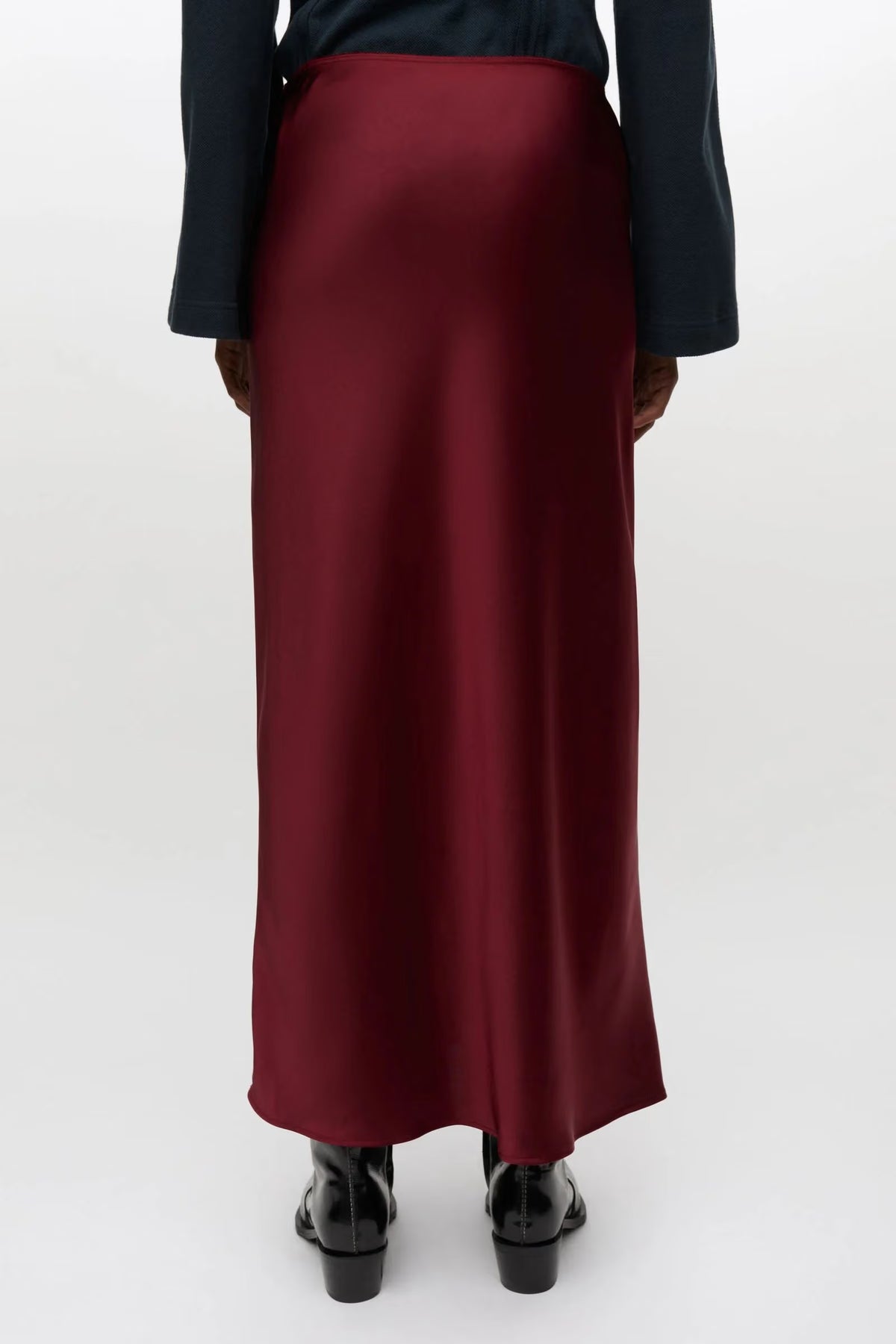 A person shown from the back wearing a long satin-like burgundy skirt with a dark navy top and black shoes, photographed against a plain light gray studio background.