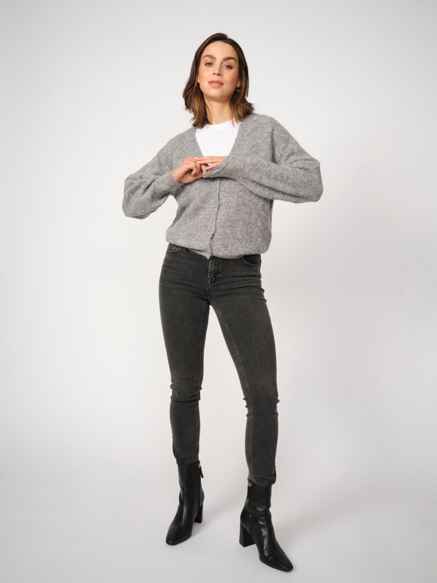 Woman wearing a gray sweater, white shirt, grey jeans, and black boots on a white background
