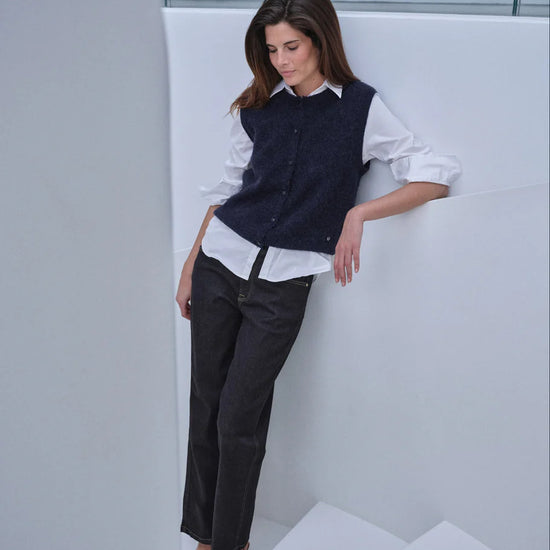 A woman models a dark navy blue knit vest over a white button-down shirt, paired with dark charcoal trousers and black pointed-toe heels, leaning against a modern white architectural wall.