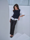 A woman models a dark navy blue knit vest over a white button-down shirt, paired with dark charcoal trousers and black pointed-toe heels, leaning against a modern white architectural wall.