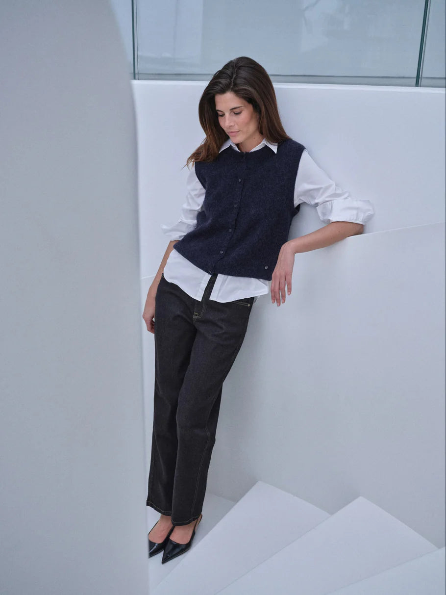 A woman models a dark navy blue knit vest over a white button-down shirt, paired with dark charcoal trousers and black pointed-toe heels, leaning against a modern white architectural wall.