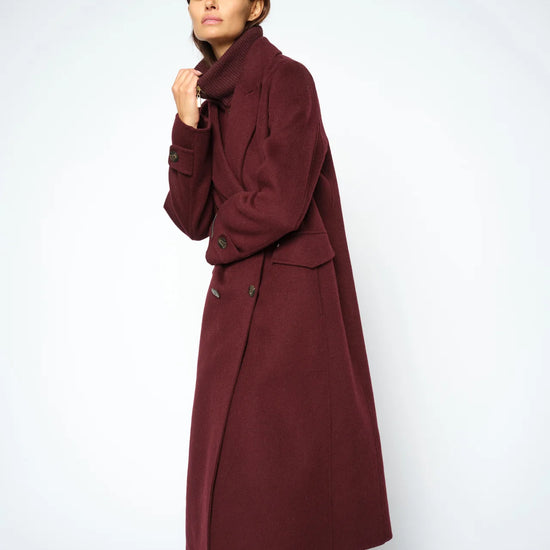A woman modeling a long, double-breasted coat in a deep burgundy color, paired with blue jeans, against a plain light gray studio background.