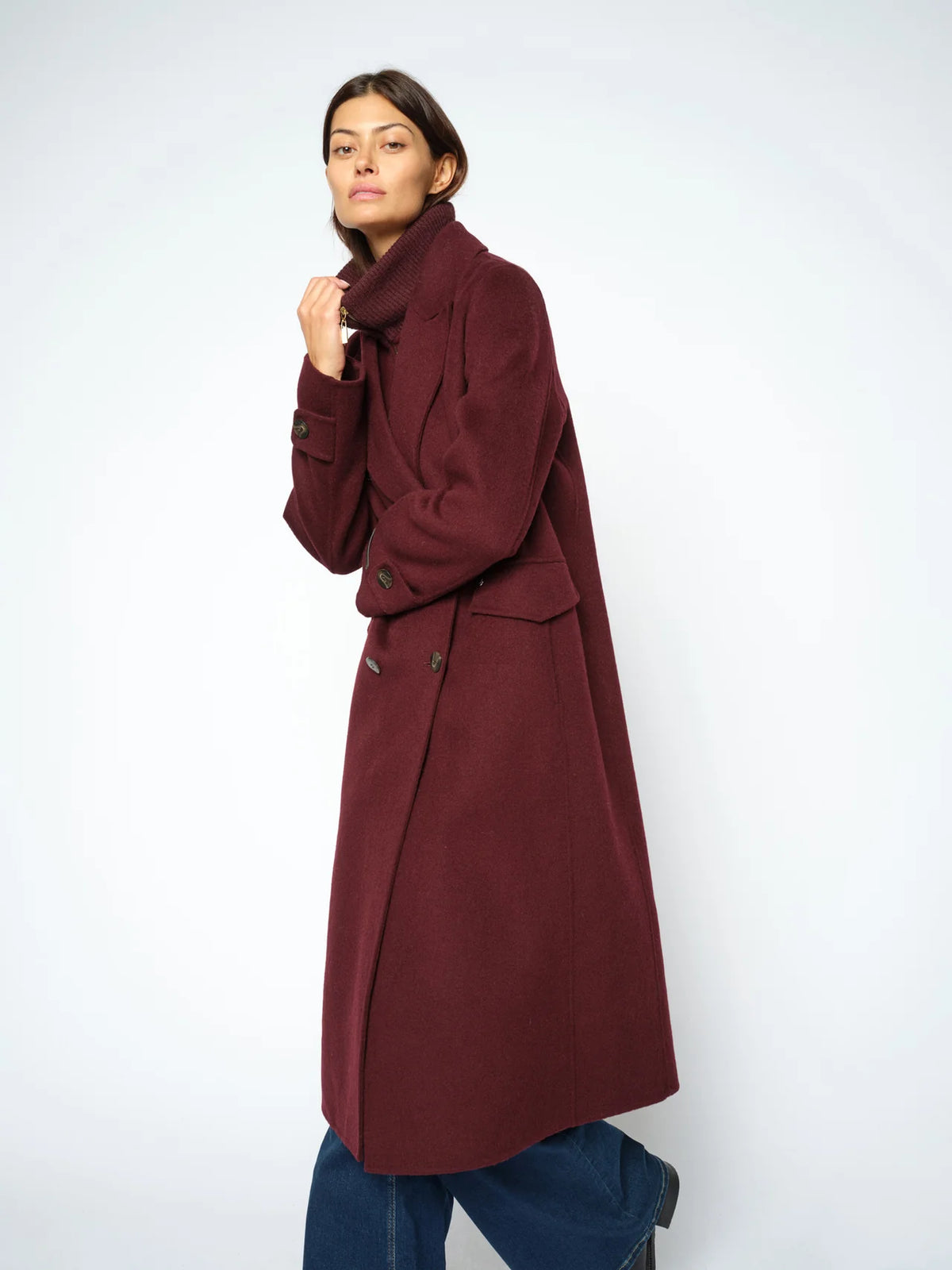 A woman modeling a long, double-breasted coat in a deep burgundy color, paired with blue jeans, against a plain light gray studio background.
