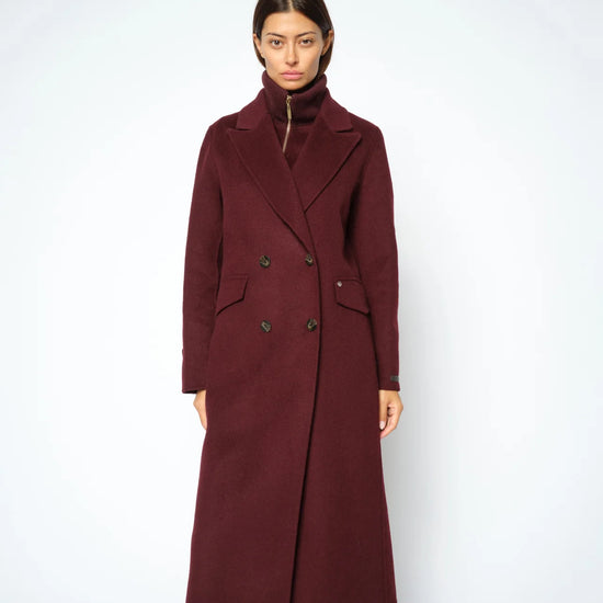 A woman modeling a long, double-breasted wool-blend coat in deep burgundy, paired with dark blue jeans, against a plain light gray studio background.