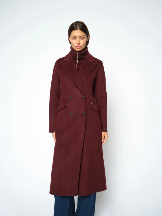 A woman modeling a long, double-breasted wool-blend coat in deep burgundy, paired with dark blue jeans, against a plain light gray studio background.