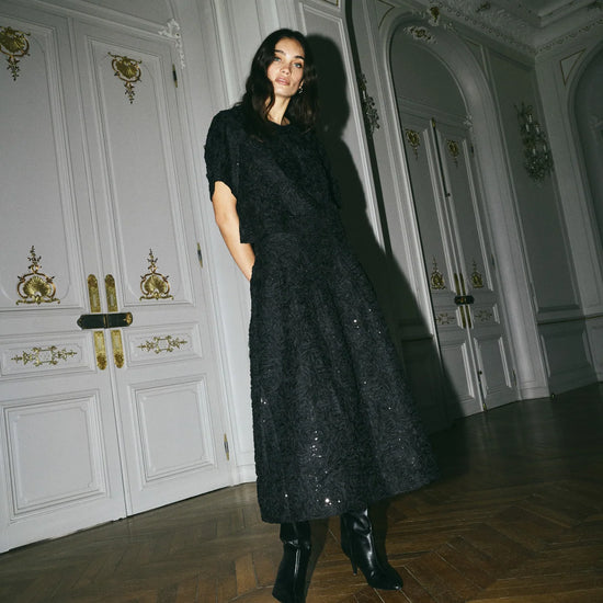 Model shown wearing a black skirt and top set with sequins on, in a grand room. 