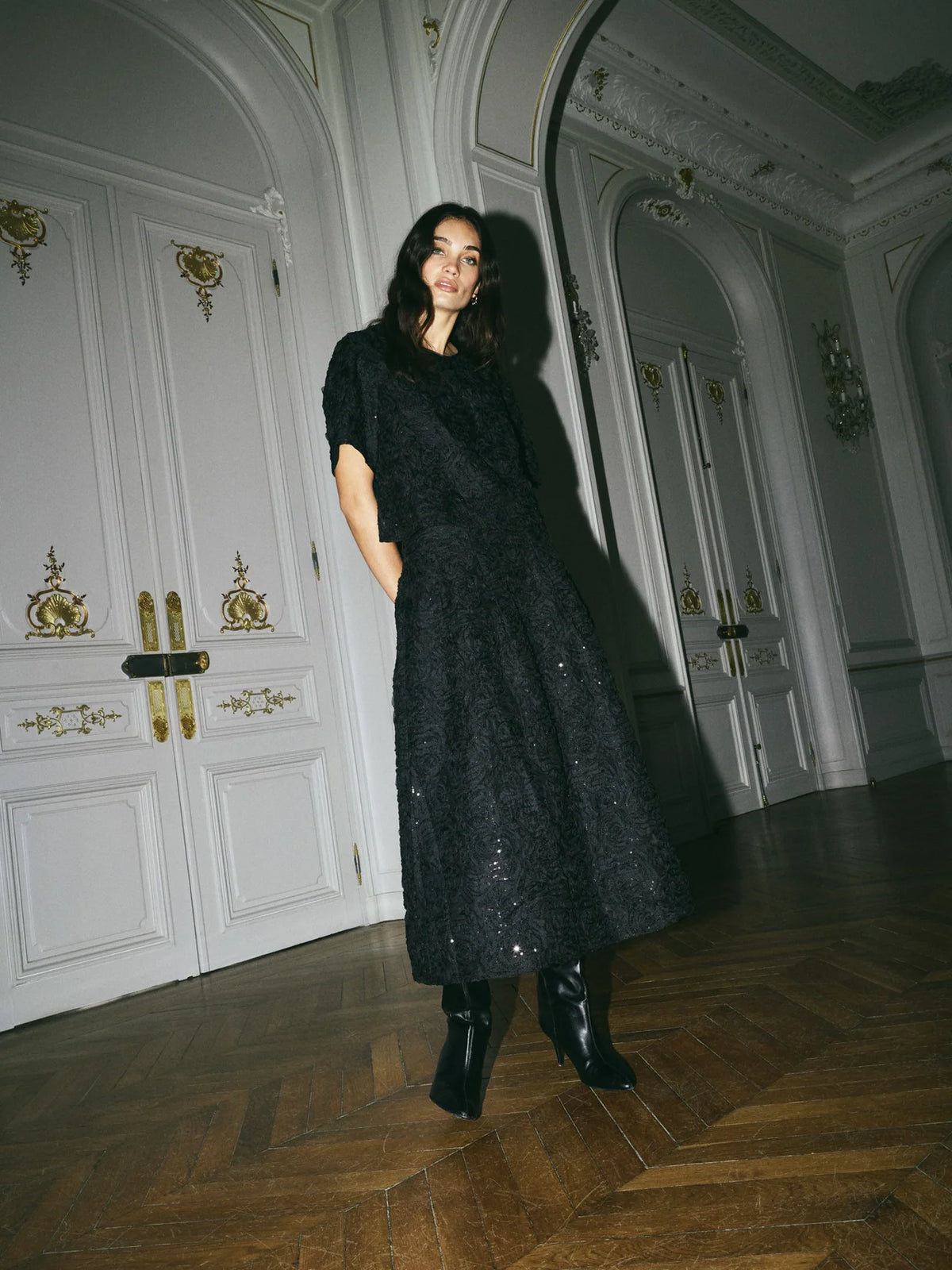 Model shown wearing a black skirt and top set with sequins on, in a grand room. 