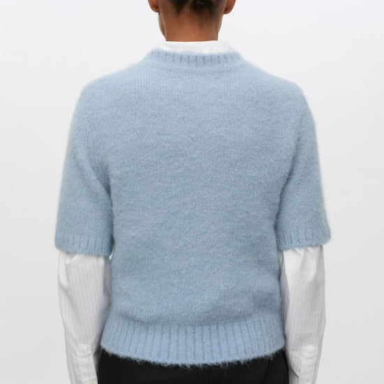 A person shown from the back wearing a light blue fuzzy short-sleeve sweater over a white collared shirt and black trousers, against a plain light background.