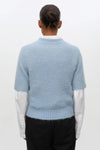 A person shown from the back wearing a light blue fuzzy short-sleeve sweater over a white collared shirt and black trousers, against a plain light background.