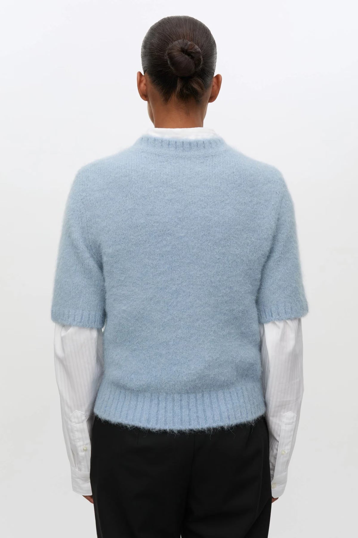 A person shown from the back wearing a light blue fuzzy short-sleeve sweater over a white collared shirt and black trousers, against a plain light background.