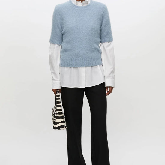 A model stands against a plain white studio background wearing a light blue knit sweater over a white collared shirt, black trousers, and black shoes, while holding a small black-and-white zebra-print handbag.