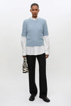 A model stands against a plain white studio background wearing a light blue knit sweater over a white collared shirt, black trousers, and black shoes, while holding a small black-and-white zebra-print handbag.