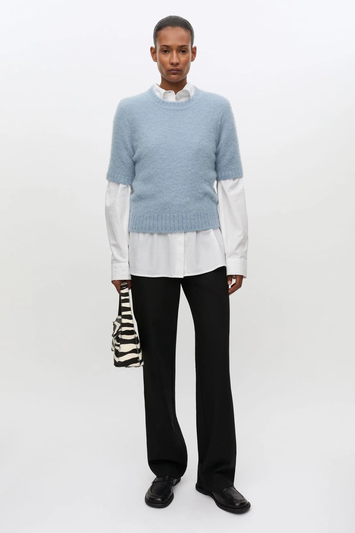 A model stands against a plain white studio background wearing a light blue knit sweater over a white collared shirt, black trousers, and black shoes, while holding a small black-and-white zebra-print handbag.