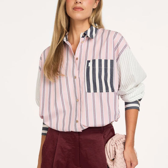 A woman modeling a pink and white vertically striped button-down shirt with navy blue striped details on the pocket and cuffs, paired with burgundy trousers and holding a light pink clutch bag.