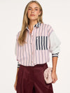 A woman modeling a pink and white vertically striped button-down shirt with navy blue striped details on the pocket and cuffs, paired with burgundy trousers and holding a light pink clutch bag.