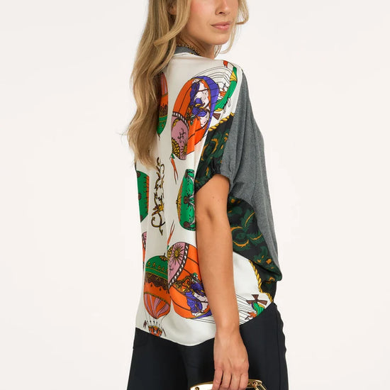 A woman modeling a short-sleeve top with a colorful graphic print on the front and a solid dark gray back panel, paired with black pants and holding a black handbag against a plain white background.