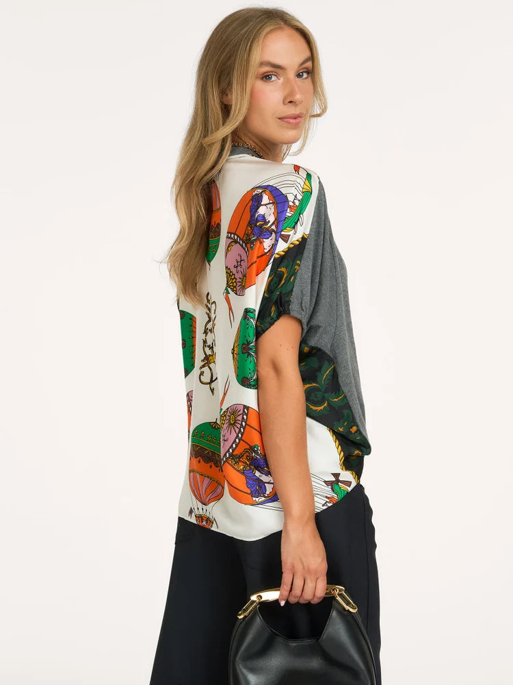 A woman modeling a short-sleeve top with a colorful graphic print on the front and a solid dark gray back panel, paired with black pants and holding a black handbag against a plain white background.