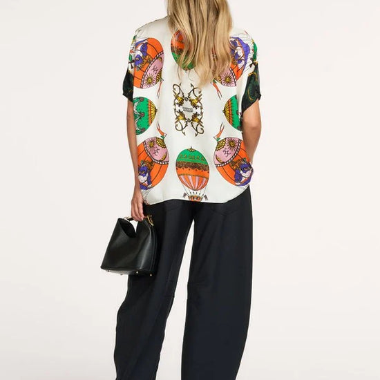 A woman shown from the back wearing a cream-colored short-sleeve top with a vibrant, multicolored graphic print of hot air balloons and a central crest, paired with wide-leg black trousers and holding a small black handbag against a plain white studio background.