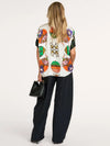 A woman shown from the back wearing a cream-colored short-sleeve top with a vibrant, multicolored graphic print of hot air balloons and a central crest, paired with wide-leg black trousers and holding a small black handbag against a plain white studio background.