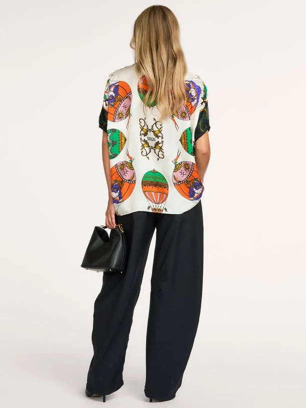 A woman shown from the back wearing a cream-colored short-sleeve top with a vibrant, multicolored graphic print of hot air balloons and a central crest, paired with wide-leg black trousers and holding a small black handbag against a plain white studio background.