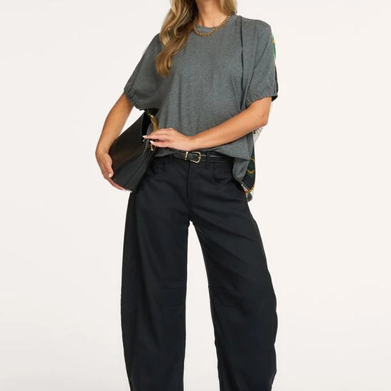 A woman modeling a gray oversized short-sleeve top, black wide-leg trousers, a black belt, and black pointed-toe heels, holding a black shoulder bag against a plain white studio background.