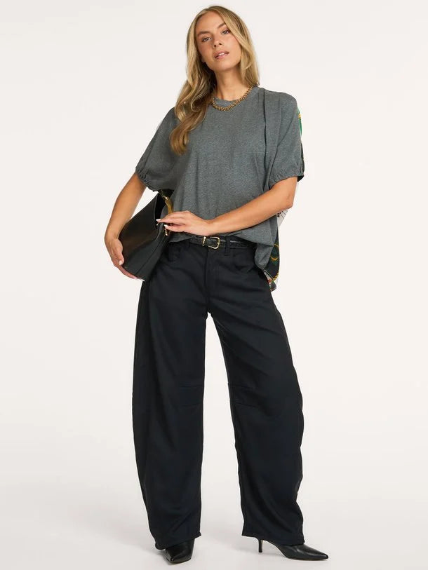 A woman modeling a gray oversized short-sleeve top, black wide-leg trousers, a black belt, and black pointed-toe heels, holding a black shoulder bag against a plain white studio background.
