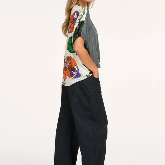 A woman modeling a short-sleeve top with a colorful butterfly print on a white base and a gray overlay, paired with wide-leg black trousers and black heels, against a plain white studio background.