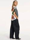 A woman modeling a short-sleeve top with a colorful butterfly print on a white base and a gray overlay, paired with wide-leg black trousers and black heels, against a plain white studio background.