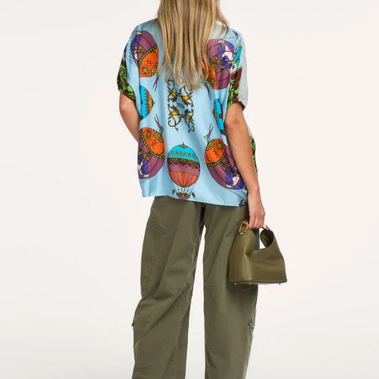 A woman shown from the back wearing a light blue short-sleeve top with a colorful retro hot air balloon print, olive green wide-leg cargo pants, and holding a small olive green handbag against a plain white studio background.