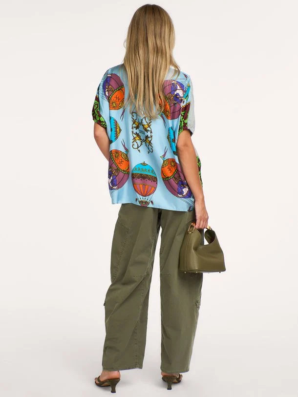 A woman shown from the back wearing a light blue short-sleeve top with a colorful retro hot air balloon print, olive green wide-leg cargo pants, and holding a small olive green handbag against a plain white studio background.